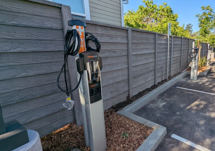 The image shows a modern electric vehicle charging station located in a parking area beside a wooden fence. The charging unit is tall and slim, equipped with multiple charging ports and cables neatly wound around holders. The area around the station is well-maintained, with mulched plant beds and clear parking lines, indicating a thoughtful integration of EV infrastructure in a residential or commercial setting.