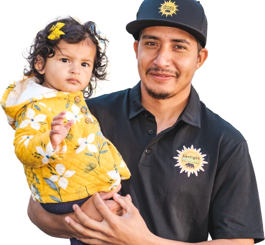 A man wearing a black polo shirt with the Sunlight Electrical logo and a matching black cap is holding a young child. The child, dressed in a bright yellow jacket adorned with white flowers, has a matching floral accessory in her hair. Both are looking directly at the camera.