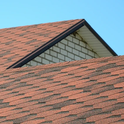 Close-up of a residential house roof with red asphalt shingles against a clear blue sky.