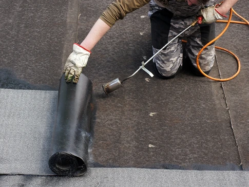 Worker using a propane torch to heat and install a black waterproofing membrane on a flat surface.